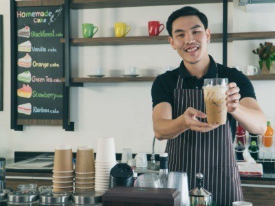 Young male student working as a barista, stood holding an iced coffee