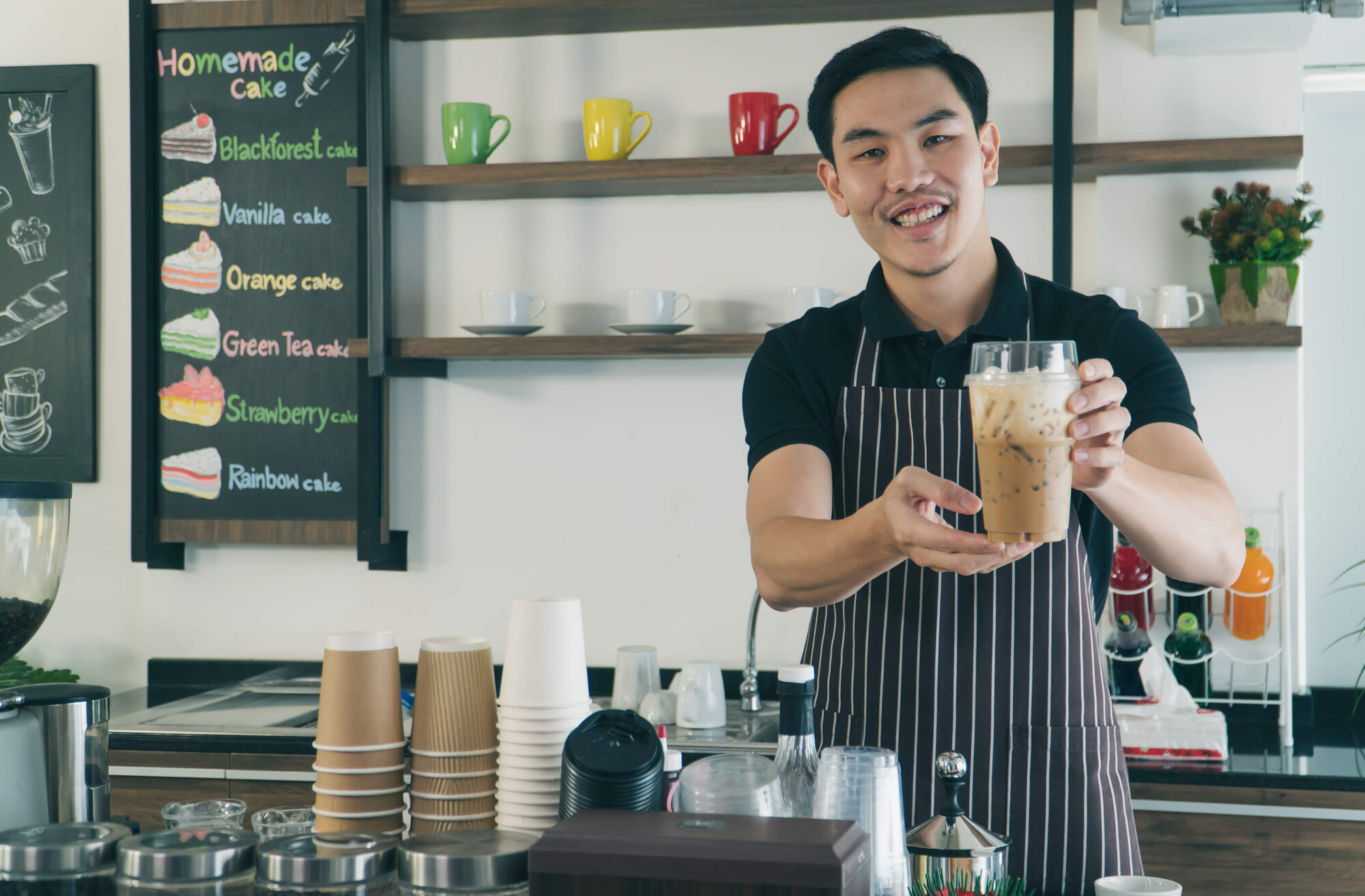 Young male student working as a barista, stood holding an iced coffee