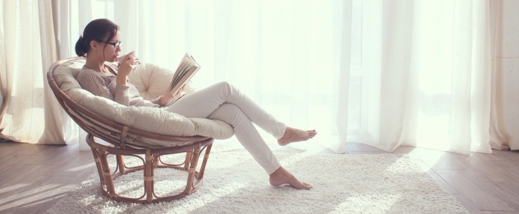 Young woman at home sitting on modern chair in front of window relaxing in her living room reading book and drinking coffee or tea
