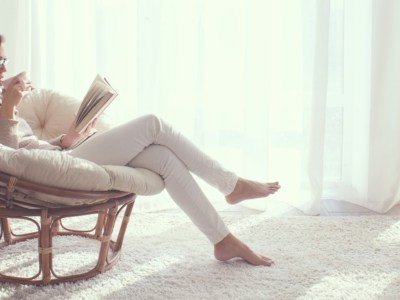 Young woman at home sitting on modern chair in front of window relaxing in her living room reading book and drinking coffee or tea
