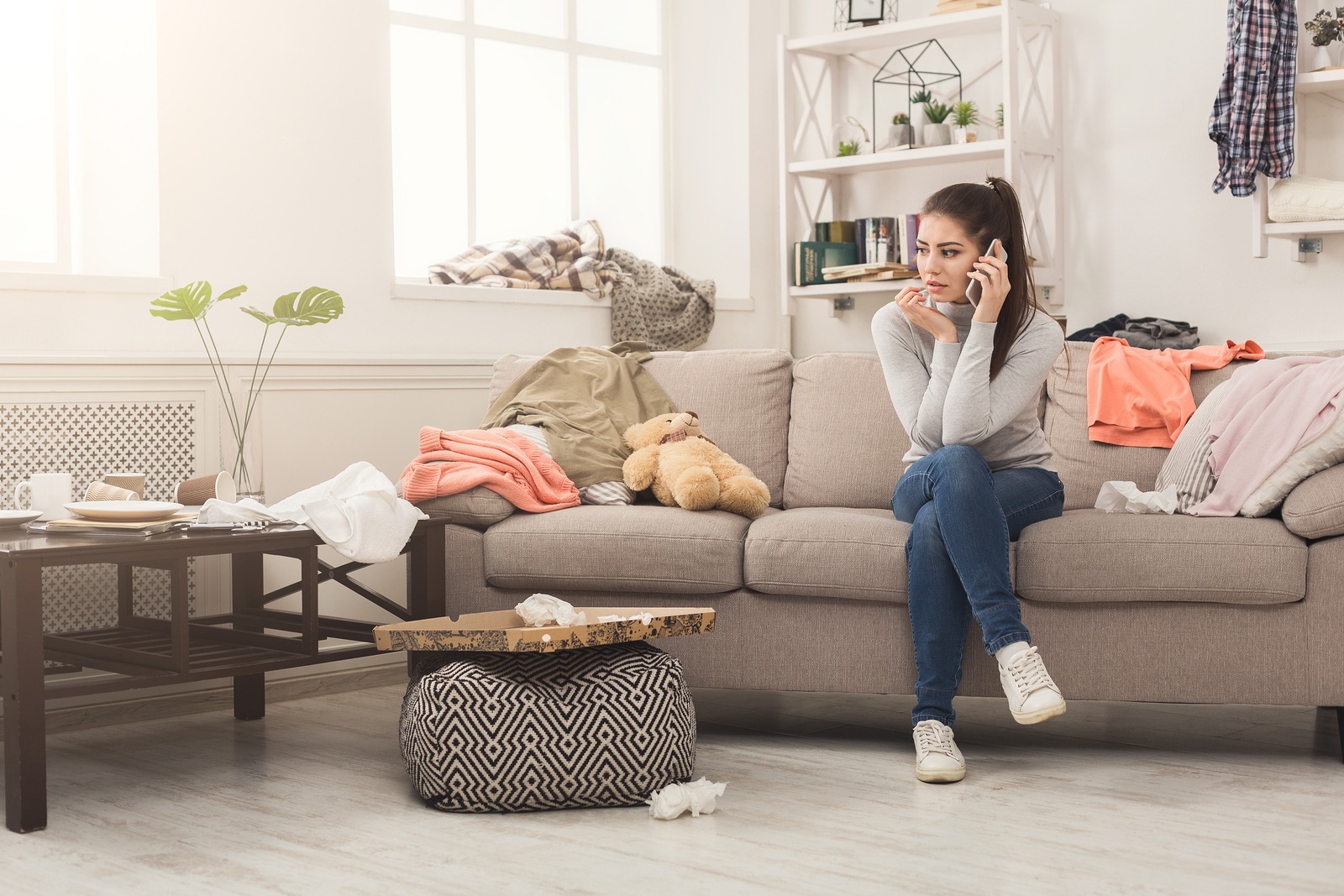 Desperate helpless woman sitting on sofa in messy living room. and talking on mobile, surrounded by many stack of clothes. Disorder and mess at home, copy space