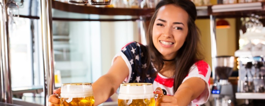 Young cheerful happy bartender girl looking at camera and offering beer to customers. Happy smiling female barista giving out beer in glasses at the bar counter. Joyful female barista is happy to be at work, having a nice day, glad to serve people concept