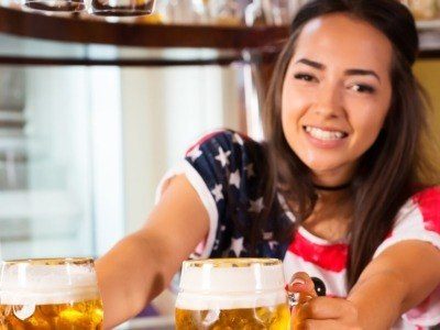 Young cheerful happy bartender girl looking at camera and offering beer to customers. Happy smiling female barista giving out beer in glasses at the bar counter. Joyful female barista is happy to be at work, having a nice day, glad to serve people concept