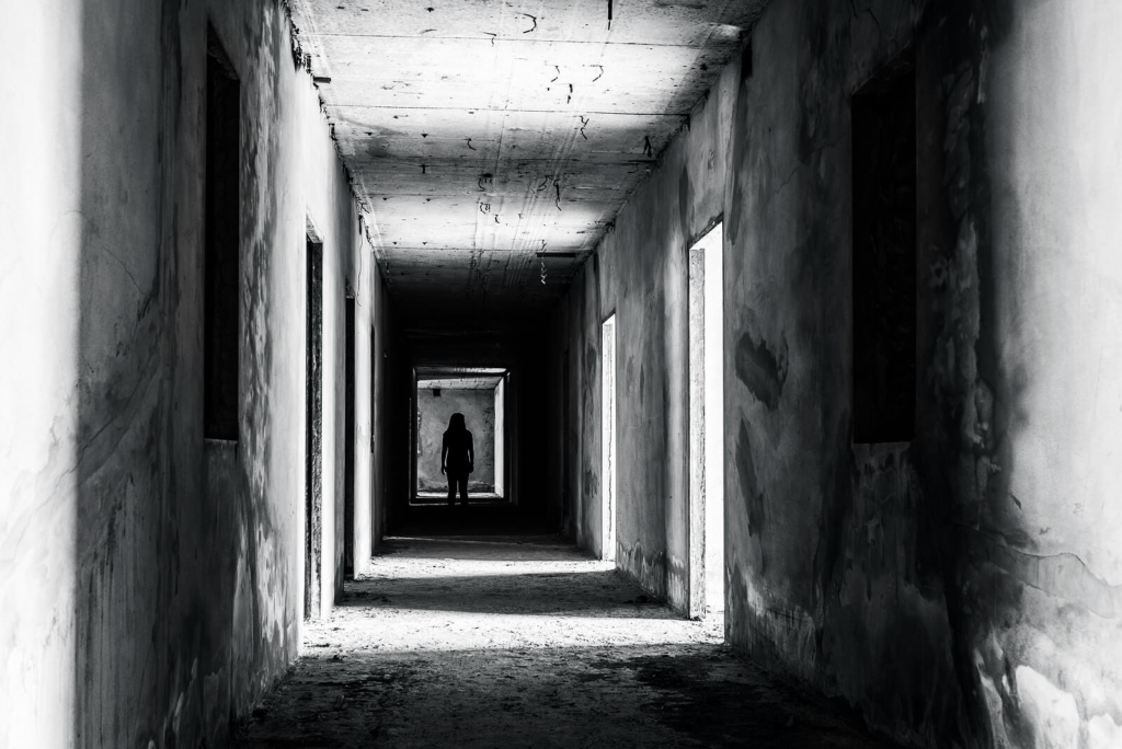 A spooky black and white corridor with a woman figure standing in the distance. Gives ghost hunt vibes.