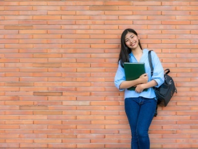 An Asian, female student leaning against a brick wall