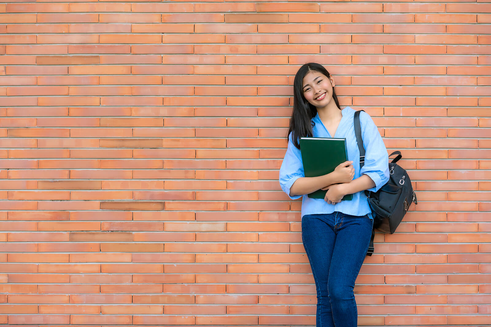 An Asian, female student leaning against a brick wall