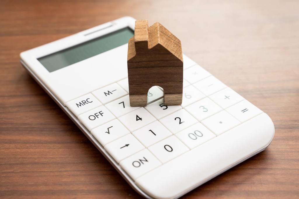 A white calculator with a wooden house shape placed on top of it, representing rent payments