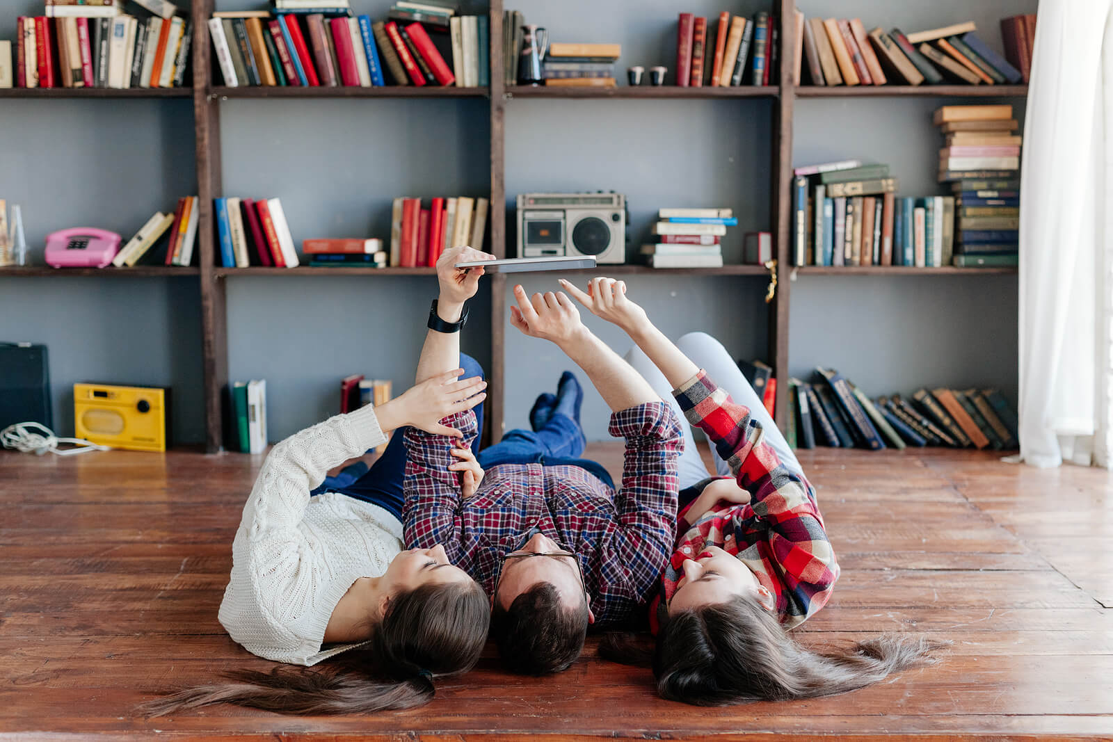 Three housemates laying on their backs on the floor of a room with lots of bookshelves, looking up at a tablet