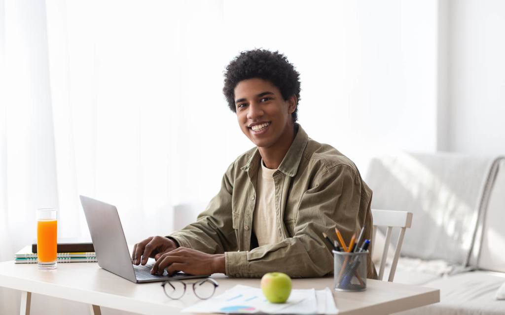 A smiling Black male student sat at a desk, typing on a laptop