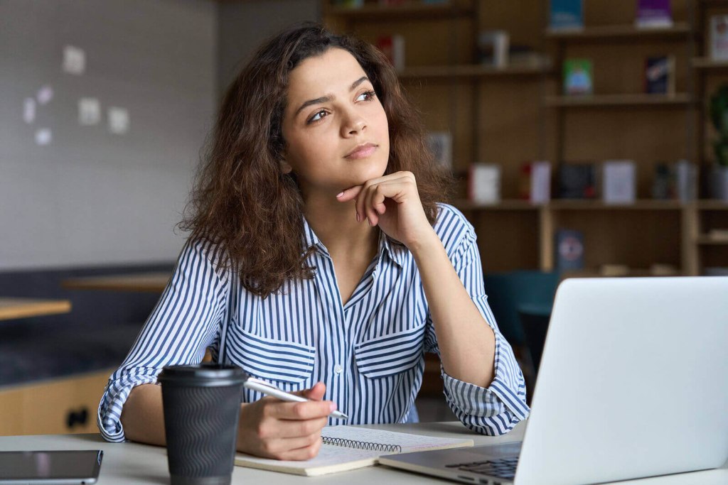 A female student wearing a blue and white stripey shirt, working on her laptop and thinking