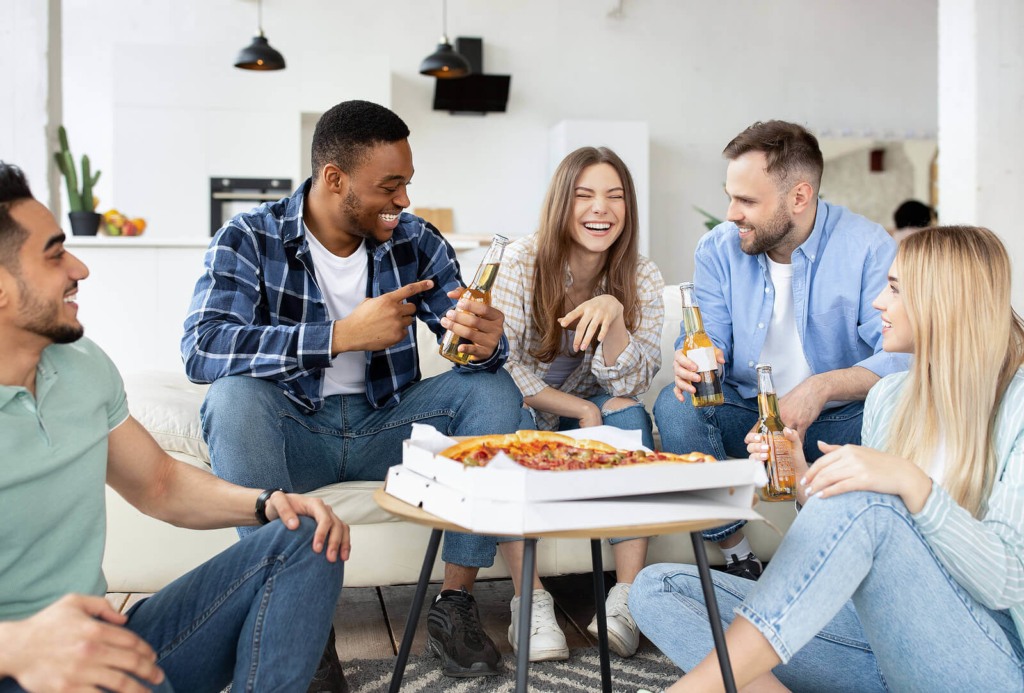 A diverse group of housemates and friends sat together in their living room eating pizza and drinking beer