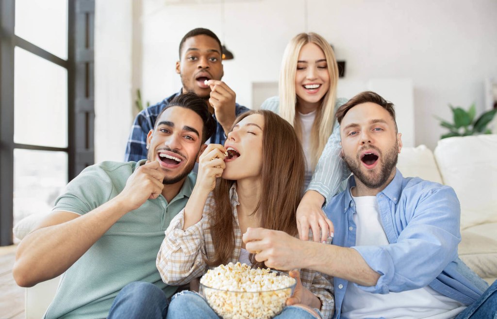 A diverse group of young people watching TV in their house share while eating popcorn