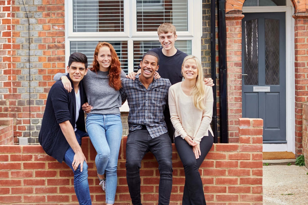 A smiling group of five students sat on a wall outside their student house share.