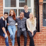 A smiling group of five students sat on a wall outside their student house share.