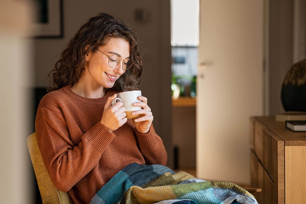 A young woman feeling cosy at home, sat enjoying a cup of tea with a blanket on her lap.