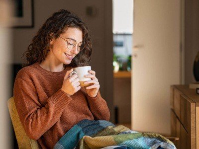 A young woman feeling cosy at home, sat enjoying a cup of tea with a blanket on her lap.