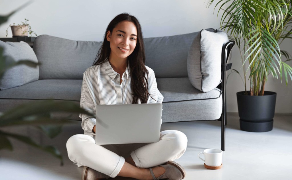 A female Asian student sat on the floor in the living room with her laptop.