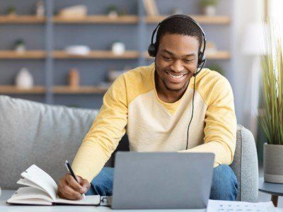 Positive black guy attending online training from home, sitting on couch in front of coffee table with laptop, using headset, taking notes, copy space.
