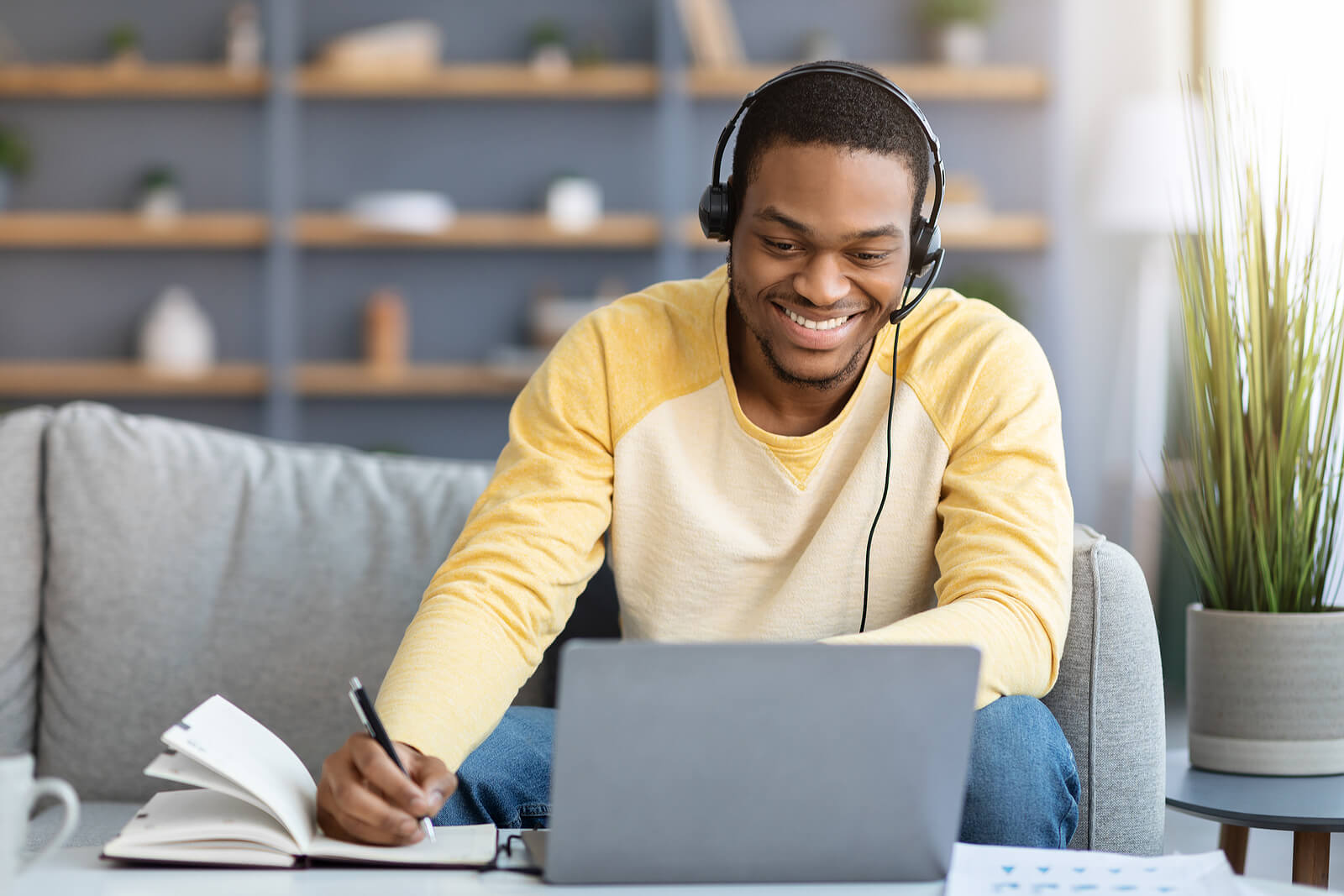 Positive black guy attending online training from home, sitting on couch in front of coffee table with laptop, using headset, taking notes, copy space.