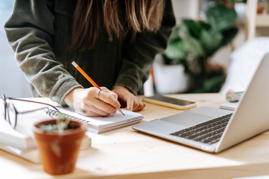 Distance learning online education and work. Business woman with plants writes in notebook. Cropped freelance girl with laptop at home office. Using computer and online shops.