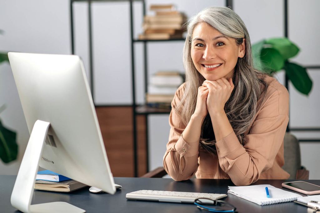 Successful influential attractive middle aged Asian female, business woman, manager, freelancer, in stylish clothes, works at computer, rests her head with her hands, looks at camera, smiles friendly

