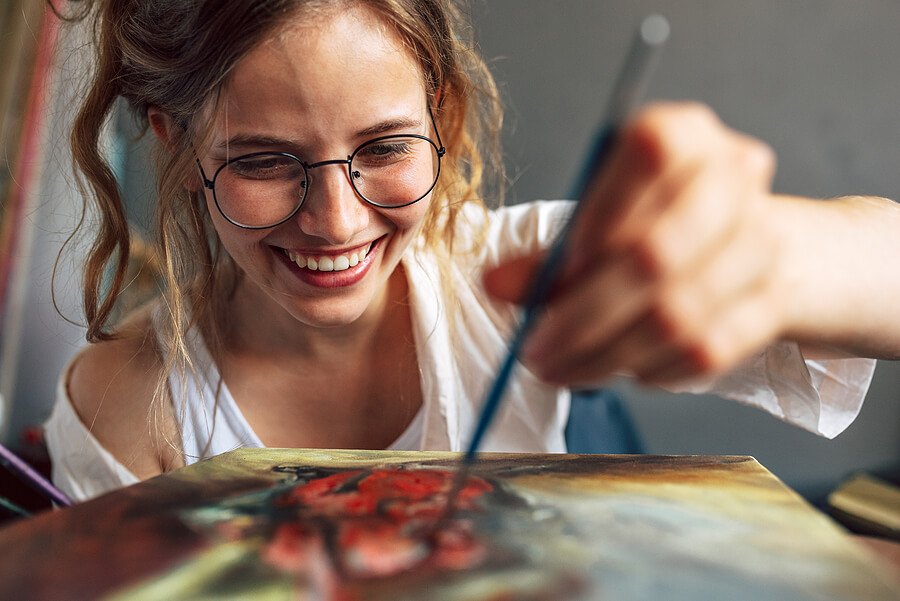 Closeup of a pretty female artist painting with a brush on canvas in her art studio. A woman painter with eyeglasses painting with oil searching for imagination in the workshop place.
