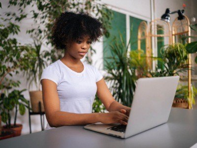 A Black woman wearing a white t-shirt, typing on her laptop at a desk surrounded by plants.