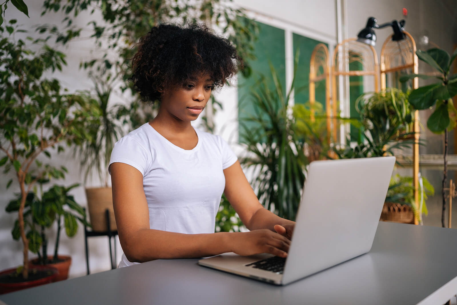 A Black woman wearing a white t-shirt, typing on her laptop at a desk surrounded by plants.