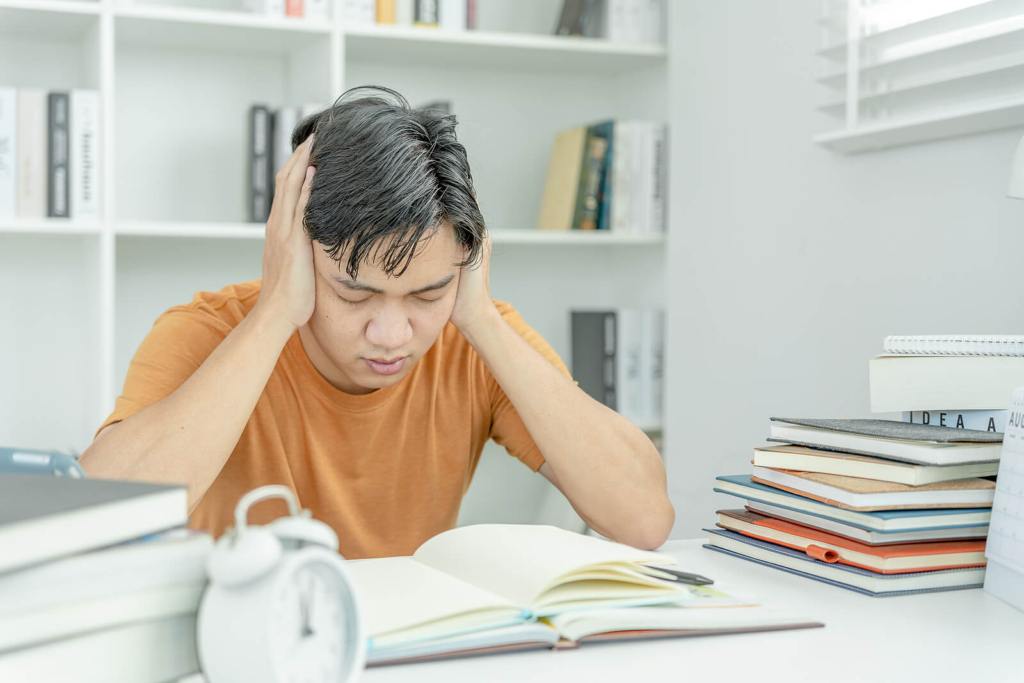 A very stressed Asian student sat at a desk covered in books for exam revision.
