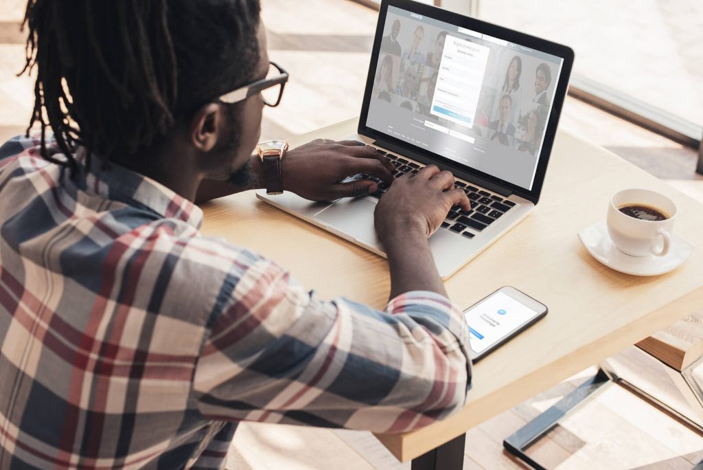 A black man in a checked shirt using LinkedIn on his laptop with a coffee and mobile phone nearby.