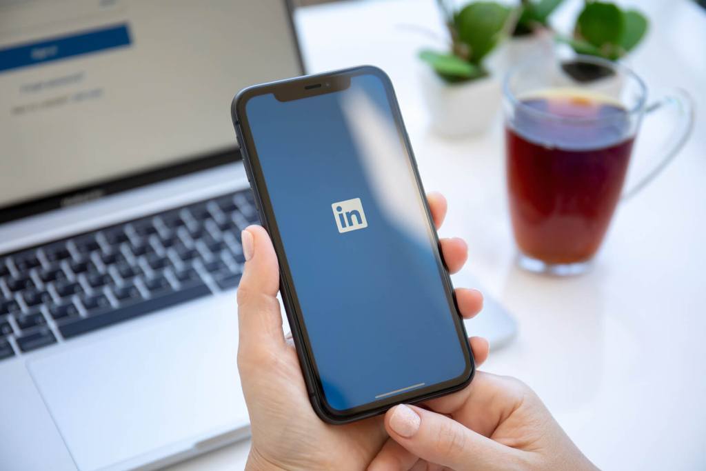 Two hands holding a mobile phone with the LinkedIn app open on the screen, while there's a laptop and coffee in the background.