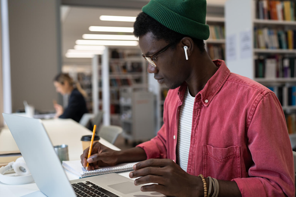 A Black man studying hard for his PhD, working on his thesis in the library with his laptop and notepad.