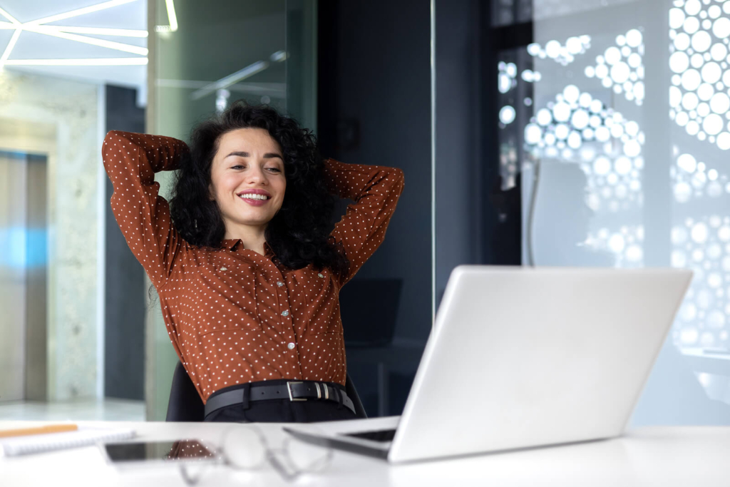 A happy woman in the office, sat at her desk and smiling at her laptop.