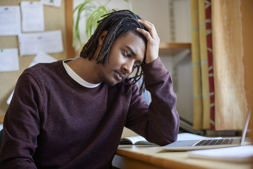 A depressed Black male university student struggling with his mental health, sat at a desk with his head in his hand.
