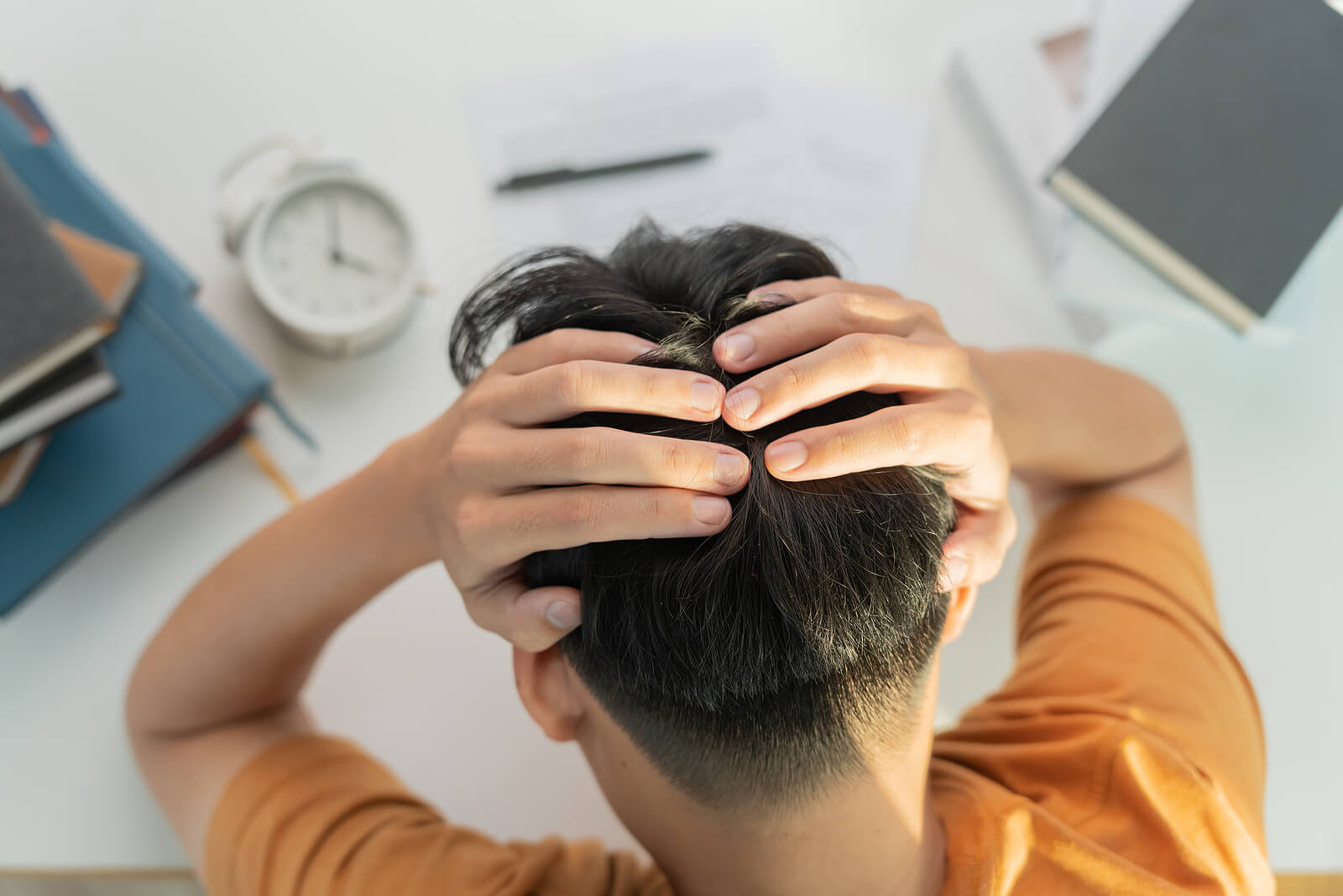 A stressed male student sat at a desk with his head in his hands. Men's mental health concept image.