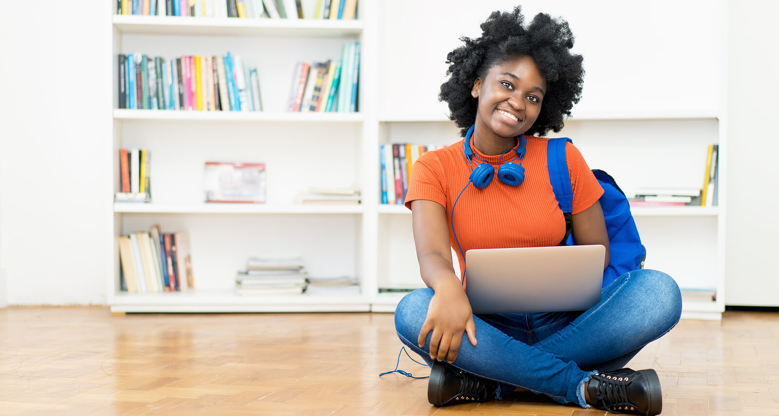 A happy, smiling Black female student sat on the floor with a laptop.