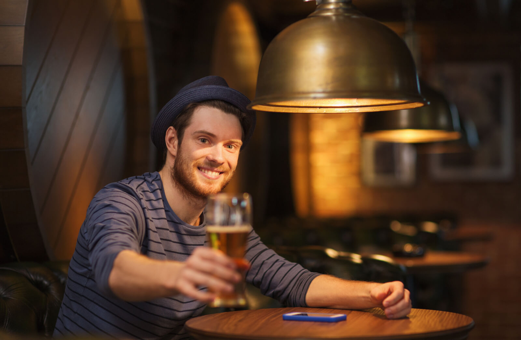 A male student drinking a pint of beer in a bar, wearing a blue hat.