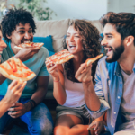 Four students eating pizza, sat in the lounge of their house share.