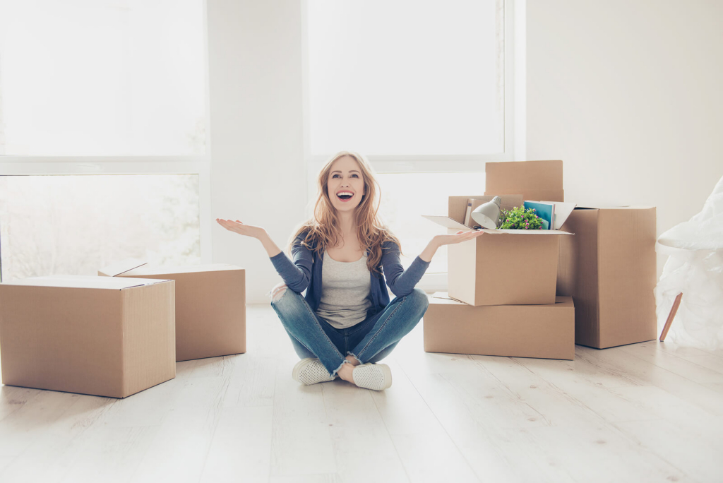 A happy woman sat on the floor of her new rental apartment, surrounded by cardboard moving boxes.