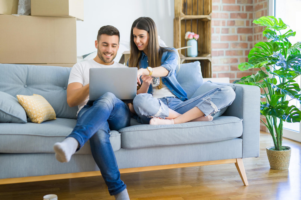 A young white couple sat on a sofa together in their first home, with moving boxes in the background. They are both looking at a laptop screen.