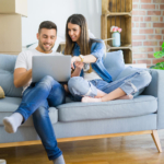 A young white couple sat on a sofa together in their first home, with moving boxes in the background. They are both looking at a laptop screen.