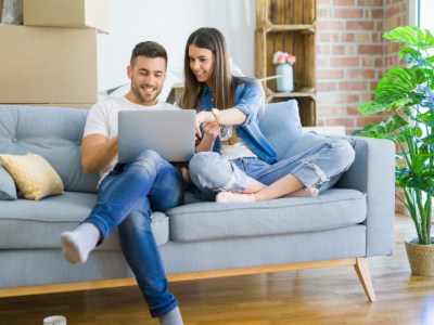 A young white couple sat on a sofa together in their first home, with moving boxes in the background. They are both looking at a laptop screen.